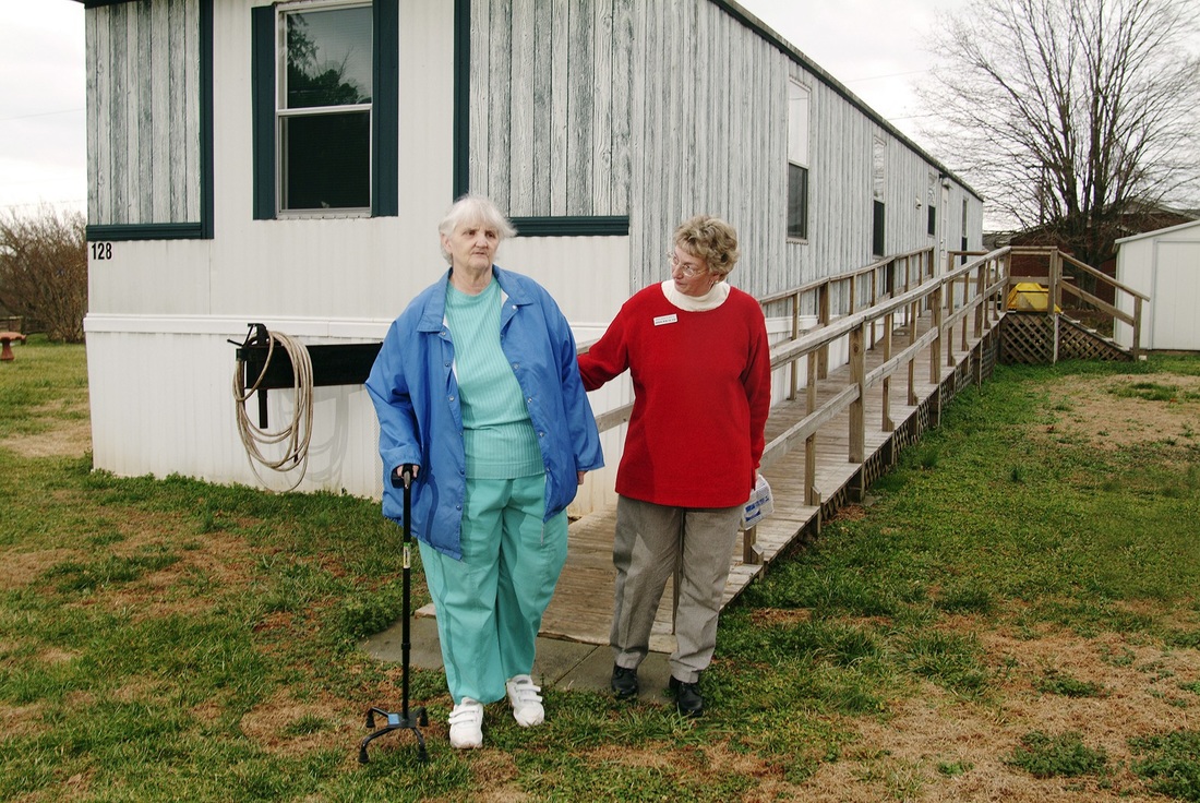 A volunteer holds the arm of an elderly women with a cane in front of the elderly women's house.