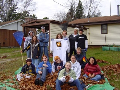 a group of children standing around and  sitting in a big pile of leaves  they raked from and elderly persons yard.
